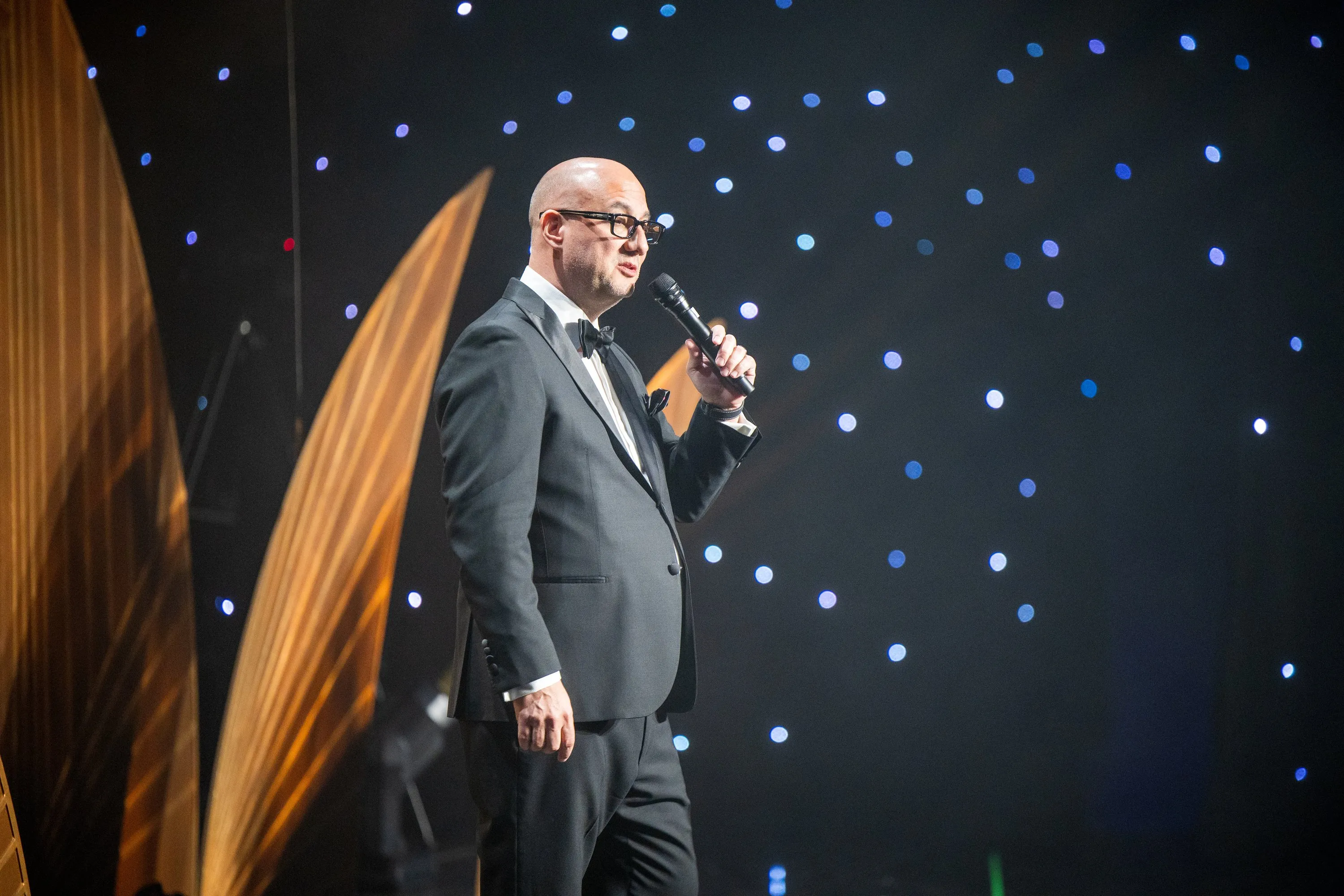Luis Giraldo at the microphone, Beverly Hilton stage, in tuxedo, blue starfield backdrop