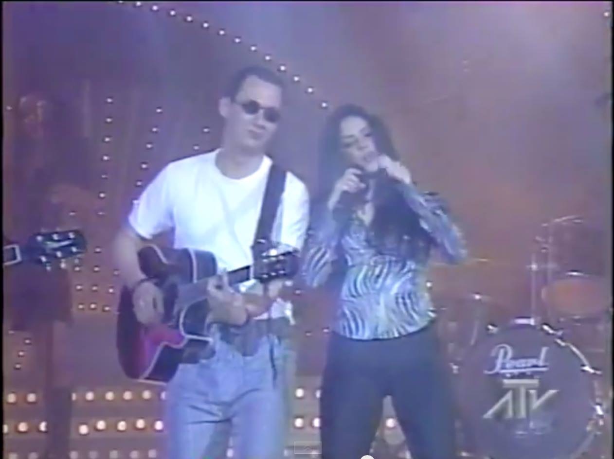 A young Luis Giraldo playing acoustic guitar alongside Shakira singing into a microphone, on a brightly-lit stage during a televised performance